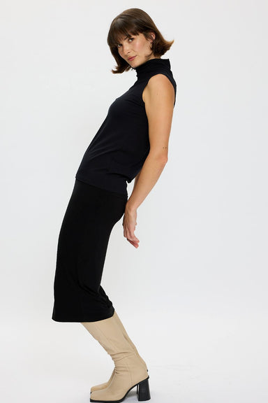 Side view of model wearing a Black sleeveless, polo neck Voyager Polo Tank with a matching Voyager midi length Skirt and paired with beige boots.